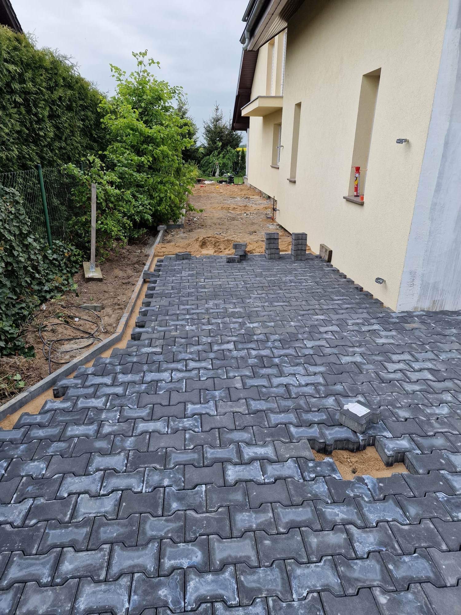 Układanie szarej kostki brukowej typu 'I' na podsypce piaskowej, widoczne krawężniki i fragment budynku w tle. Częściowo ułożona nawierzchnia.