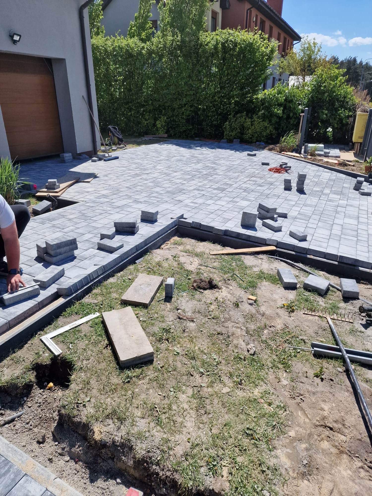 Fragment układania szarej kostki brukowej na podjeździe domu jednorodzinnego. Widoczne narzędzia i częściowo ułożona nawierzchnia. Prace w toku, dzień słoneczny.