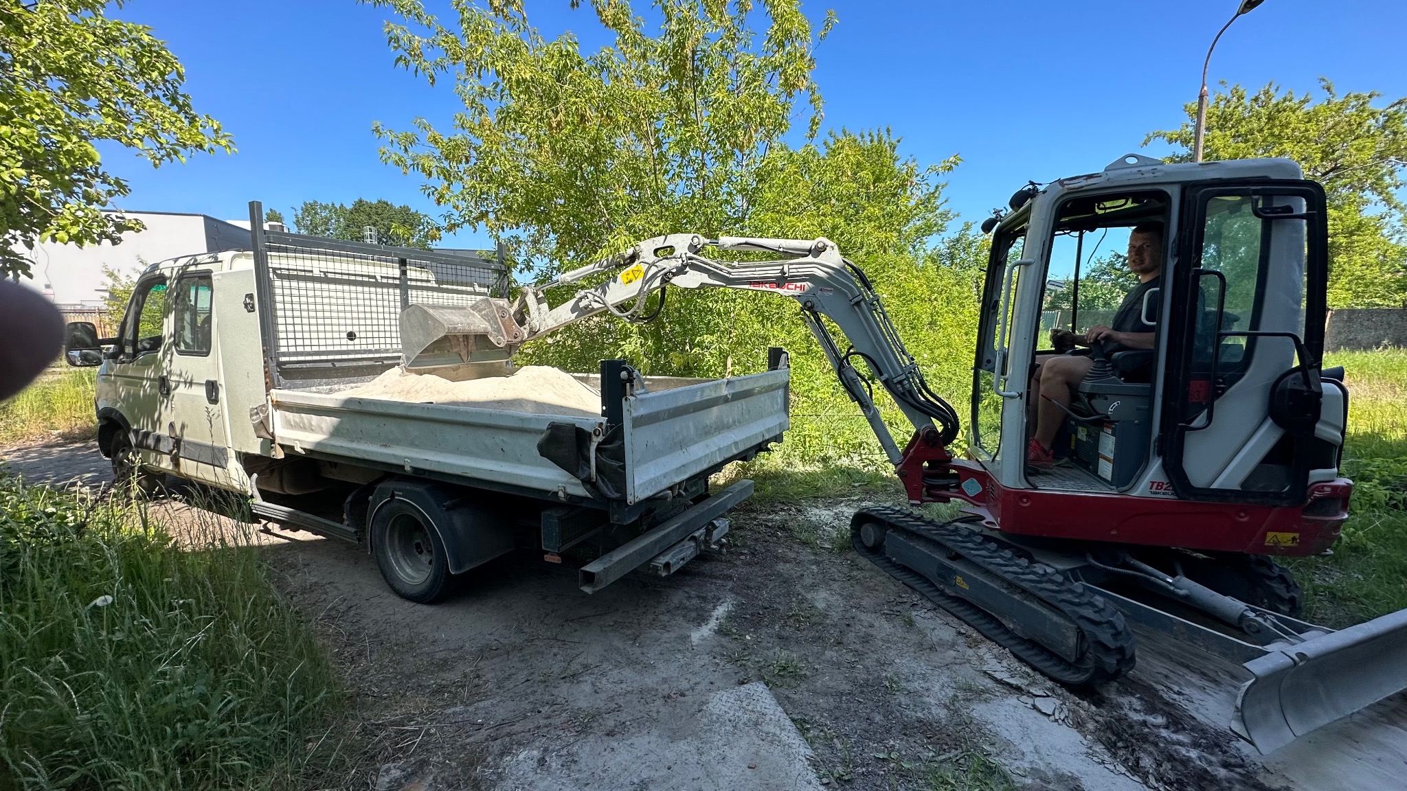 Mini koparka ładuje piasek do skrzyni samochodu ciężarowego na tle zieleni. Operator koparki w kabinie. Praca w toku, słoneczny dzień.