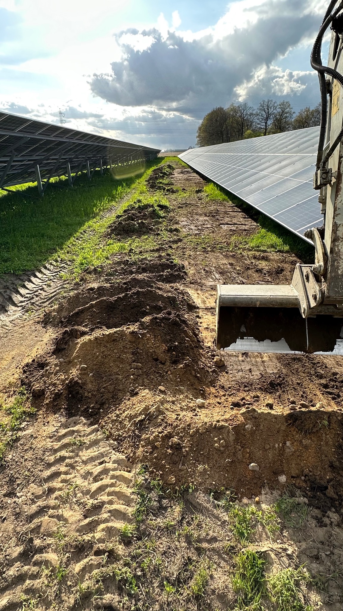 Wykop pod panele słoneczne wykonany koparką na terenie farmy fotowoltaicznej. Ślady opon na ziemi, pochmurne niebo w tle. Prace ziemne w toku.