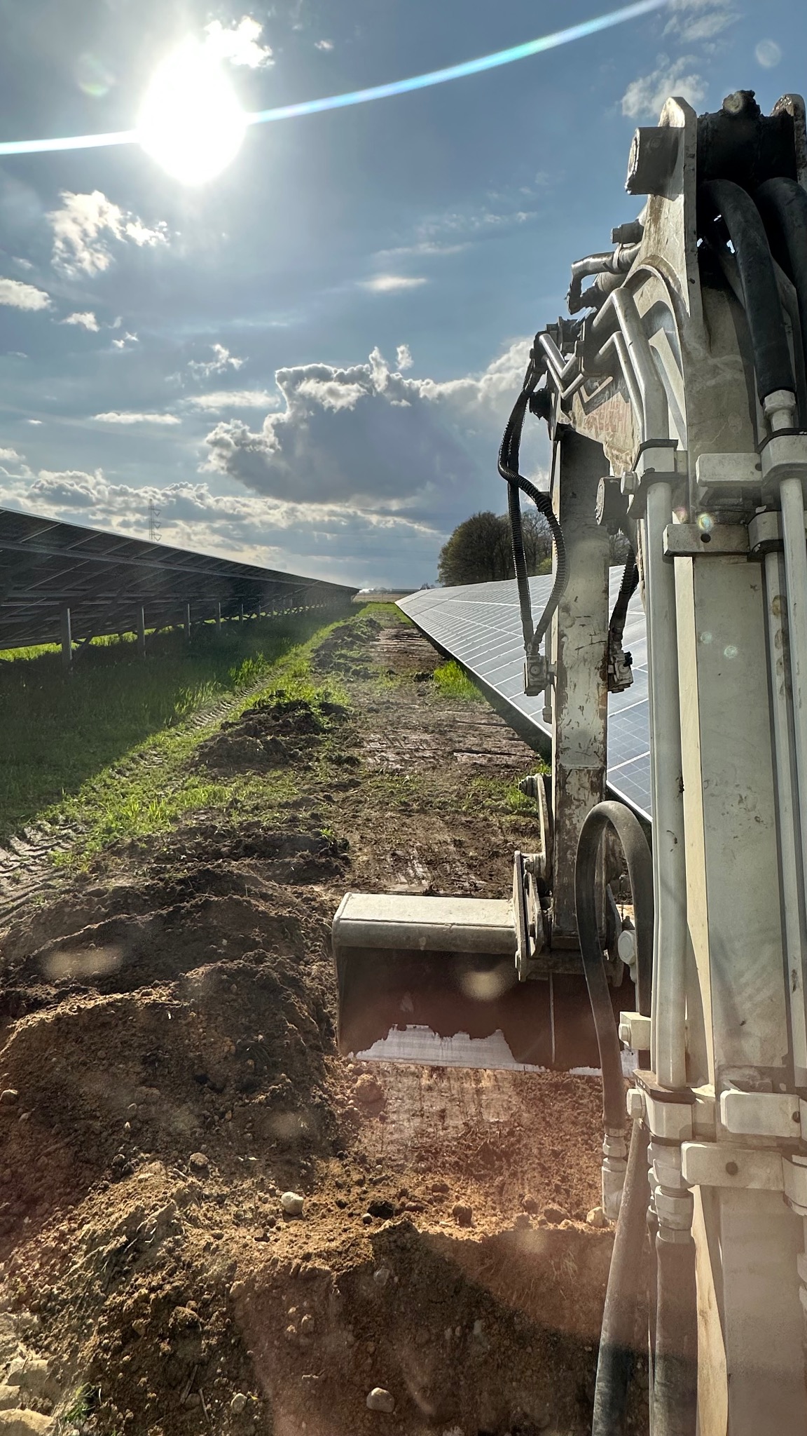 Koparka w trakcie prac ziemnych przy panelach słonecznych. Widoczne ślady opon na ziemi i błękitne niebo z chmurami w tle. Prace przygotowawcze pod instalacje.