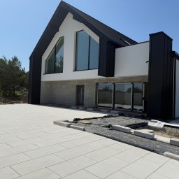 FIRMA HANDLOWO-USŁUGOWA DAMIAN GRODECKI - Nowoczesny dom z czarno-białą elewacją i dużymi oknami, w trakcie prac wykończeniowych na zewnątrz. Widoczne ułożone płyty chodnikowe i przygotowania do dalszych prac.