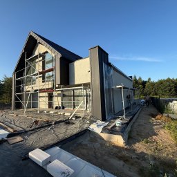 FIRMA HANDLOWO-USŁUGOWA DAMIAN GRODECKI - Nowoczesny dom w trakcie prac elewacyjnych: rusztowania, folia ochronna, przygotowania do tynkowania. Widok z zewnątrz, w słoneczny dzień.