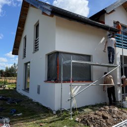 FIRMA HANDLOWO-USŁUGOWA DAMIAN GRODECKI - Biała elewacja domu w trakcie tynkowania. Ekipa na rusztowaniu, okna zabezpieczone folią, widoczne wiadra z tynkiem i narzędzia. Słoneczny dzień, błękitne niebo.
