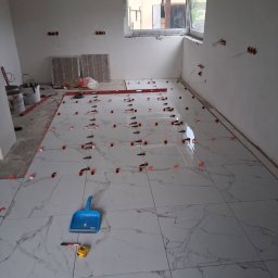 FIRMA HANDLOWO-USŁUGOWA DAMIAN GRODECKI - Układanie białych, marmurkowych płytek podłogowych z użyciem klipsów poziomujących. Widoczne narzędzia: poziomica, miarka, szufelka. Surowy stan ścian i okno w tle.