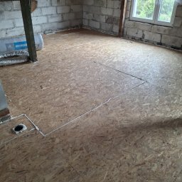 FIRMA HANDLOWO-USŁUGOWA DAMIAN GRODECKI - Płyty OSB na poddaszu, przygotowane do dalszych prac wykończeniowych. Widoczne białe uszczelnienia i izolacja. Surowy stan, okno z widokiem na zieleń.