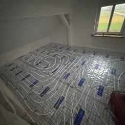 FIRMA HANDLOWO-USŁUGOWA DAMIAN GRODECKI - Białe rury ogrzewania podłogowego ułożone na siatce w szarym pomieszczeniu z oknem. Widok z góry, instalacja przed wylaniem posadzki, etap prac.