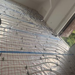 FIRMA HANDLOWO-USŁUGOWA DAMIAN GRODECKI - Białe rury ogrzewania podłogowego, przymocowane czarnymi uchwytami do siatki na izolacji, ułożone w jasnym pomieszczeniu. Widoczne narzędzia.