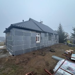FIRMA HANDLOWO-USŁUGOWA DAMIAN GRODECKI - Dom jednorodzinny w trakcie ocieplania, ściany pokryte szarymi płytami izolacyjnymi. Widoczne okna, dach z ciemnej dachówki i elementy rusztowania na posesji.