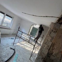 FIRMA HANDLOWO-USŁUGOWA DAMIAN GRODECKI - Pracownik na rusztowaniu skuwa fragment sufitu w jasnym pomieszczeniu z oknem. Widoczne gruz i narzędzia, co sugeruje etap remontu domu lub kamienicy.