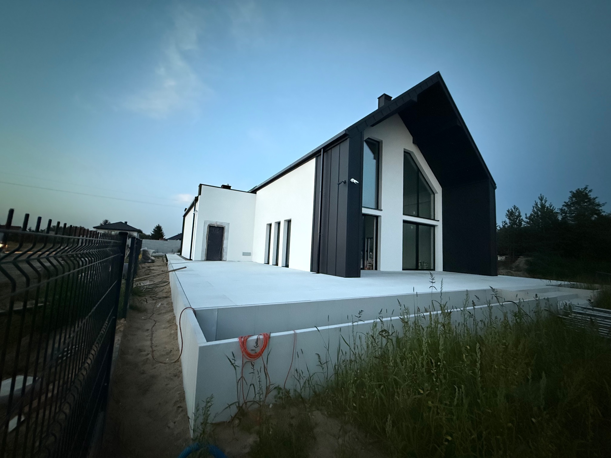 Nowoczesny dom jednorodzinny z białą elewacją i czarnymi akcentami, widoczny z zewnątrz, z elementami wykończeniowymi w trakcie realizacji.