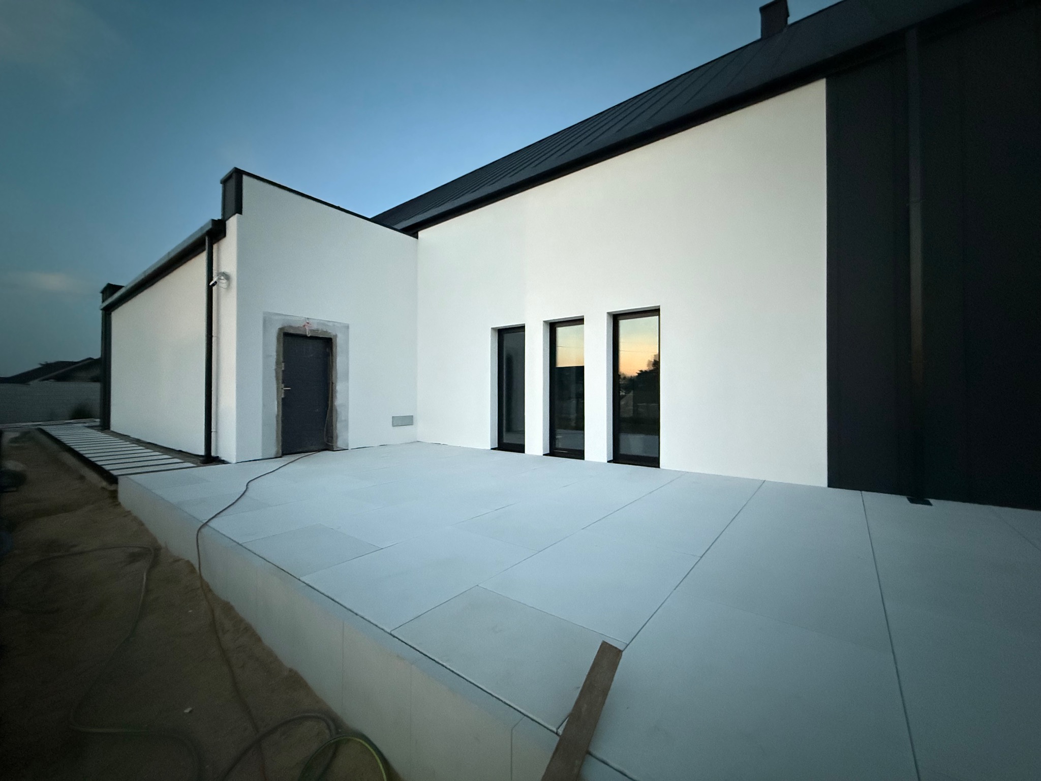 Nowoczesny dom z białą elewacją i czarnymi akcentami, widoczne trzy okna i drzwi w trakcie wykończenia. Minimalistyczny design, równomierne oświetlenie.