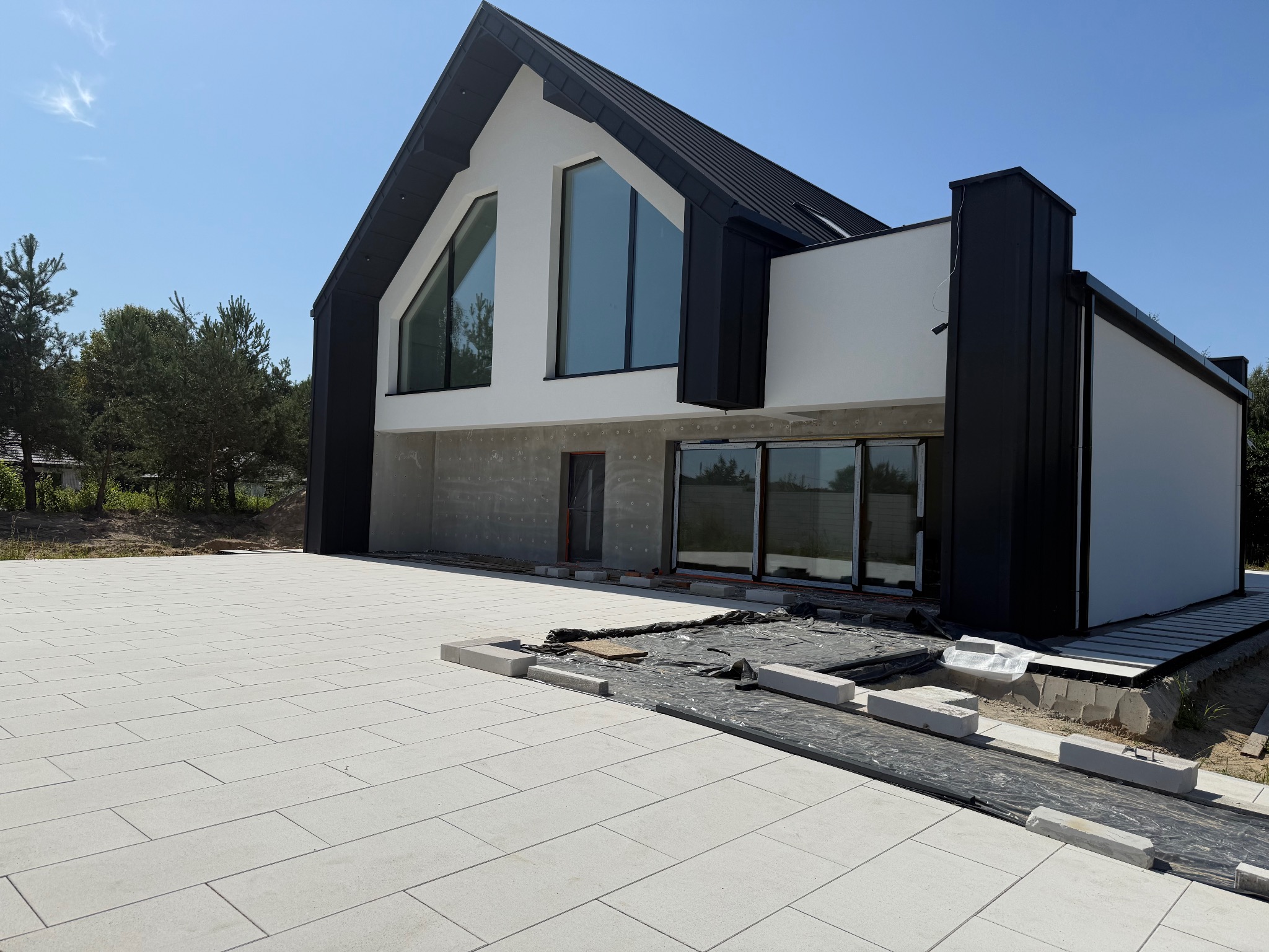 Nowoczesny dom z czarno-białą elewacją i dużymi oknami, w trakcie prac wykończeniowych na zewnątrz. Widoczne ułożone płyty chodnikowe i przygotowania do dalszych prac.