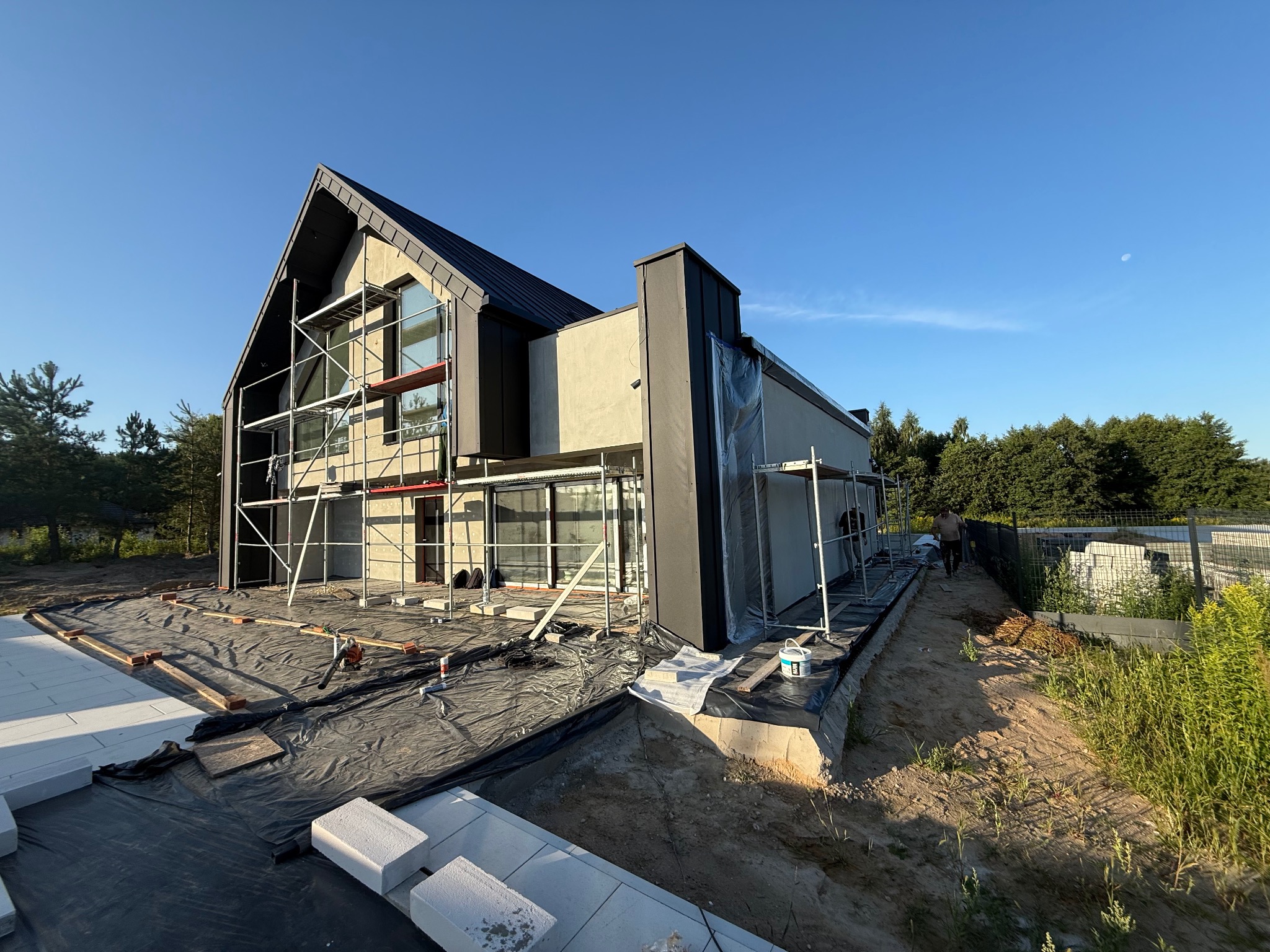 Nowoczesny dom w trakcie prac elewacyjnych: rusztowania, folia ochronna, przygotowania do tynkowania. Widok z zewnątrz, w słoneczny dzień.