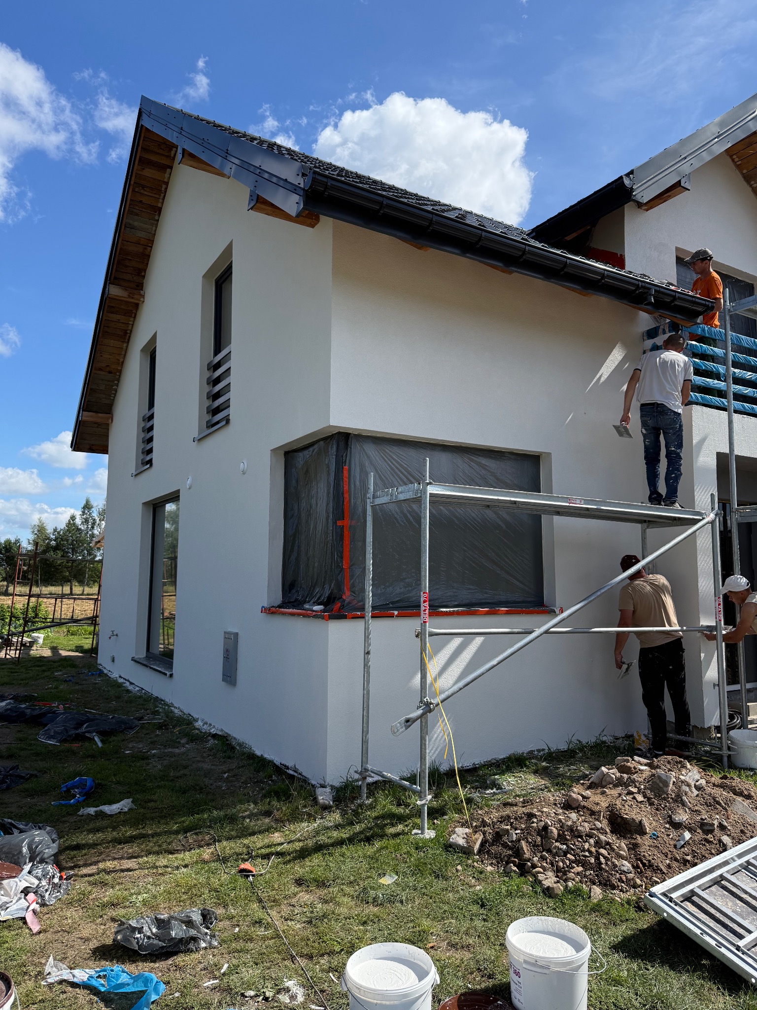 Biała elewacja domu w trakcie tynkowania. Ekipa na rusztowaniu, okna zabezpieczone folią, widoczne wiadra z tynkiem i narzędzia. Słoneczny dzień, błękitne niebo.