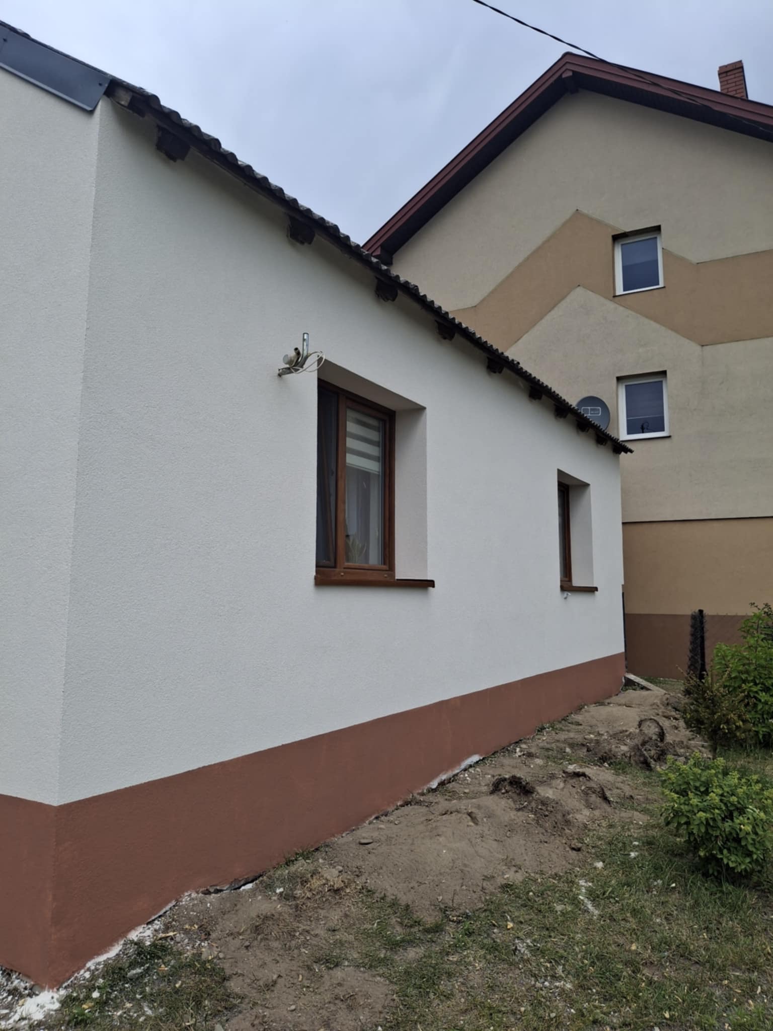 Otynkowana i pomalowana elewacja budynku z brązowym cokołem. Widoczne dwa okna z brązowymi ramami. W tle budynek z elewacją w odcieniach beżu i brązu.