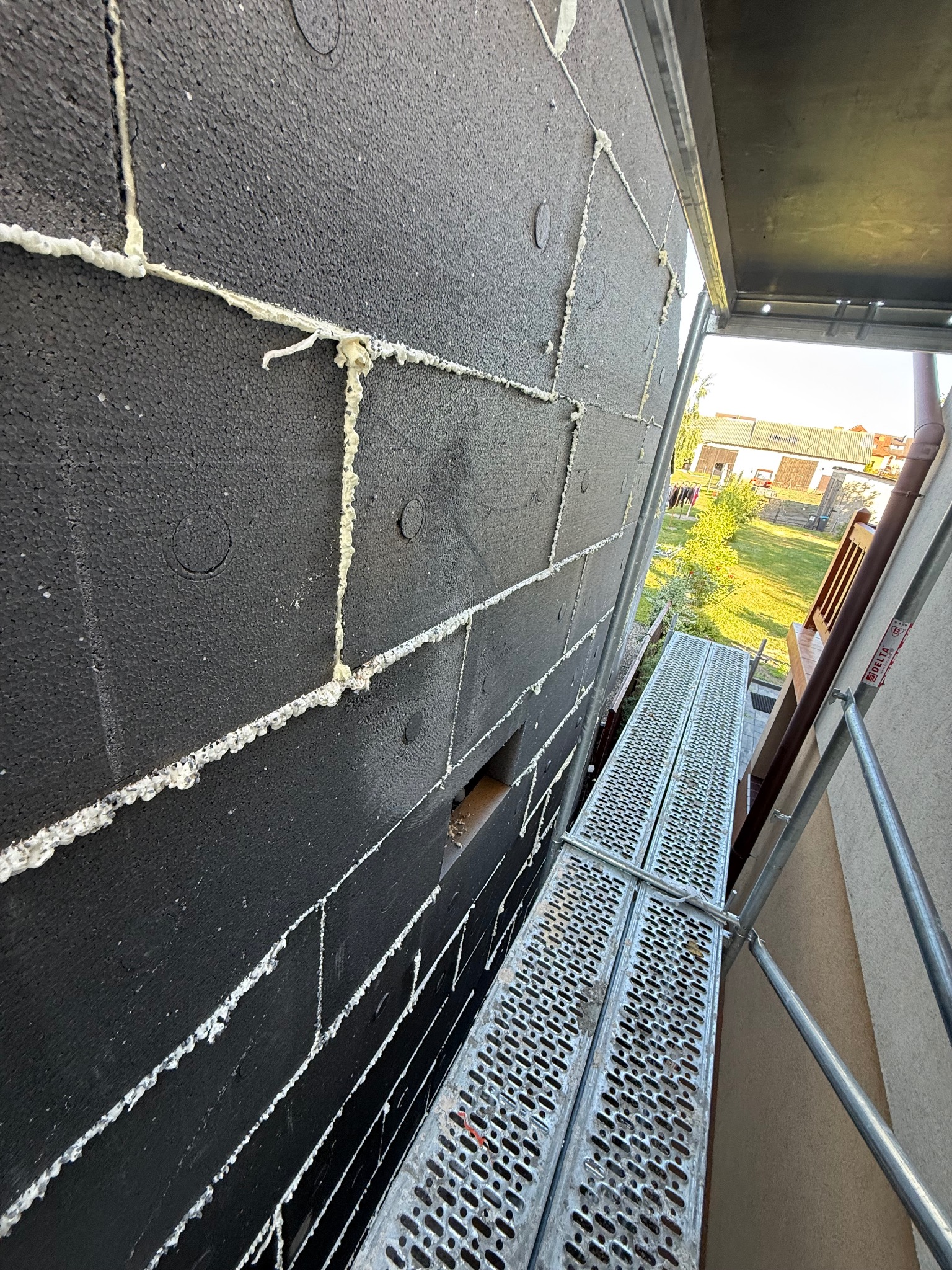 Czarny styropian na elewacji budynku w Nowej Wsi, przyklejony pianką montażową. Widok z rusztowania, z boku. Etap ocieplania.