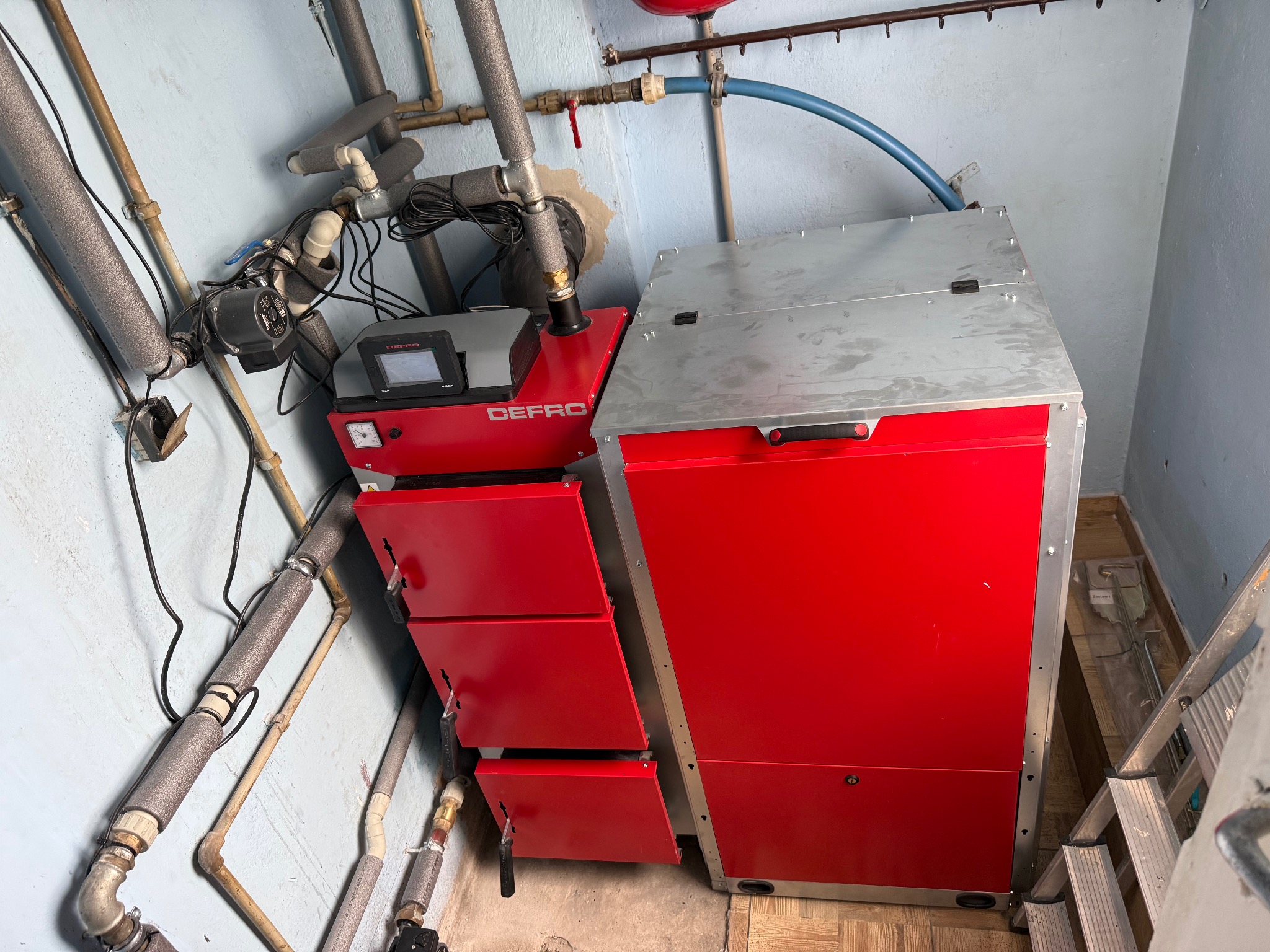 Czerwony piec DEFRO z otwartymi drzwiczkami w kotłowni, widoczne rury i instalacje. Metalowa drabina oparta o ścianę. Ujęcie z dołu, podkreślające instalację.