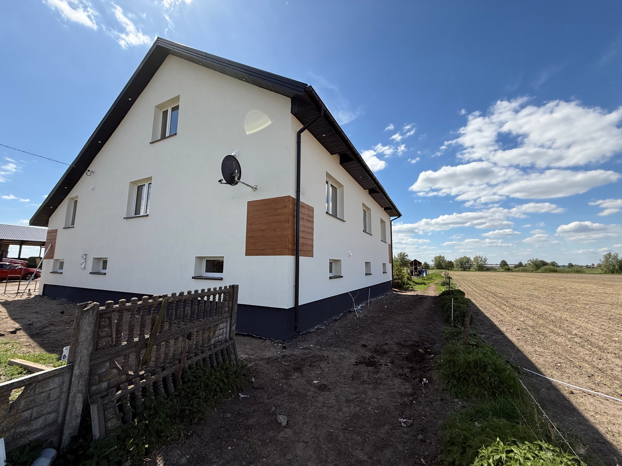 Biała elewacja domu z czarnym dachem i brązowymi akcentami, widoczna antena satelitarna, okna z białymi ramami, tło: błękitne niebo i pole.