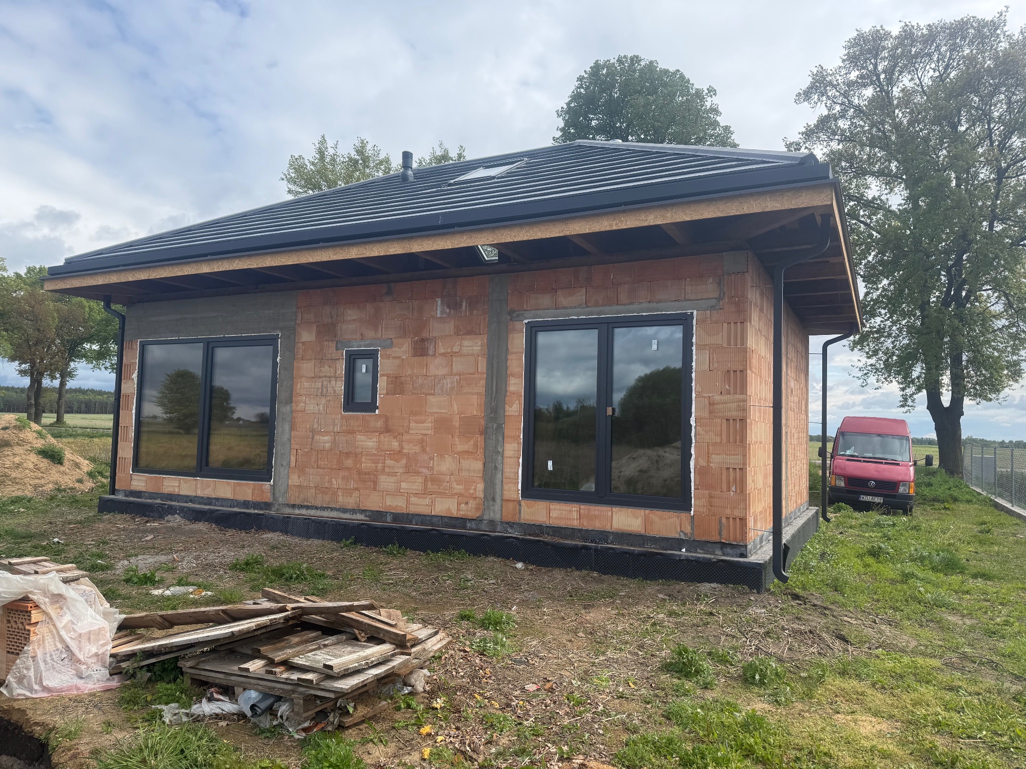 Surowy budynek z czerwonej cegły z czarnymi oknami i dachem, widoczne drewniane elementy konstrukcyjne, teren budowy z resztkami materiałów, krajobraz wiejski w tle.