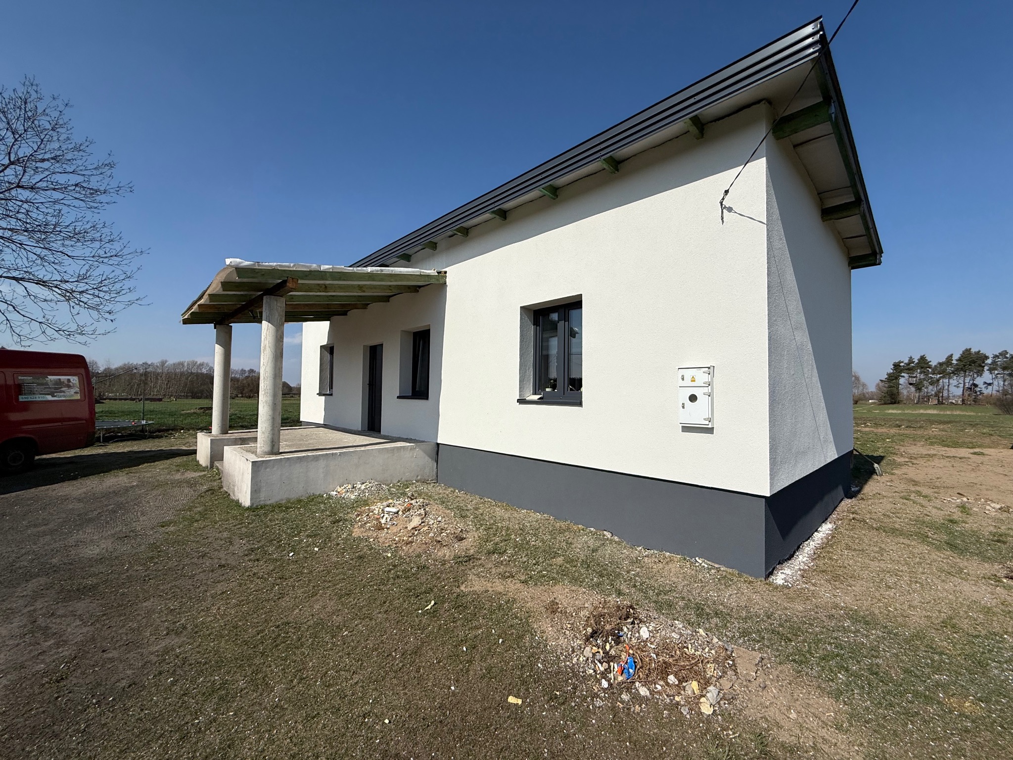 Otynkowany dom jednorodzinny z szarą podmurówką i zadaszonym wejściem. Biała elewacja w kontraście z błękitnym niebem. Widoczny fragment czerwonego samochodu.