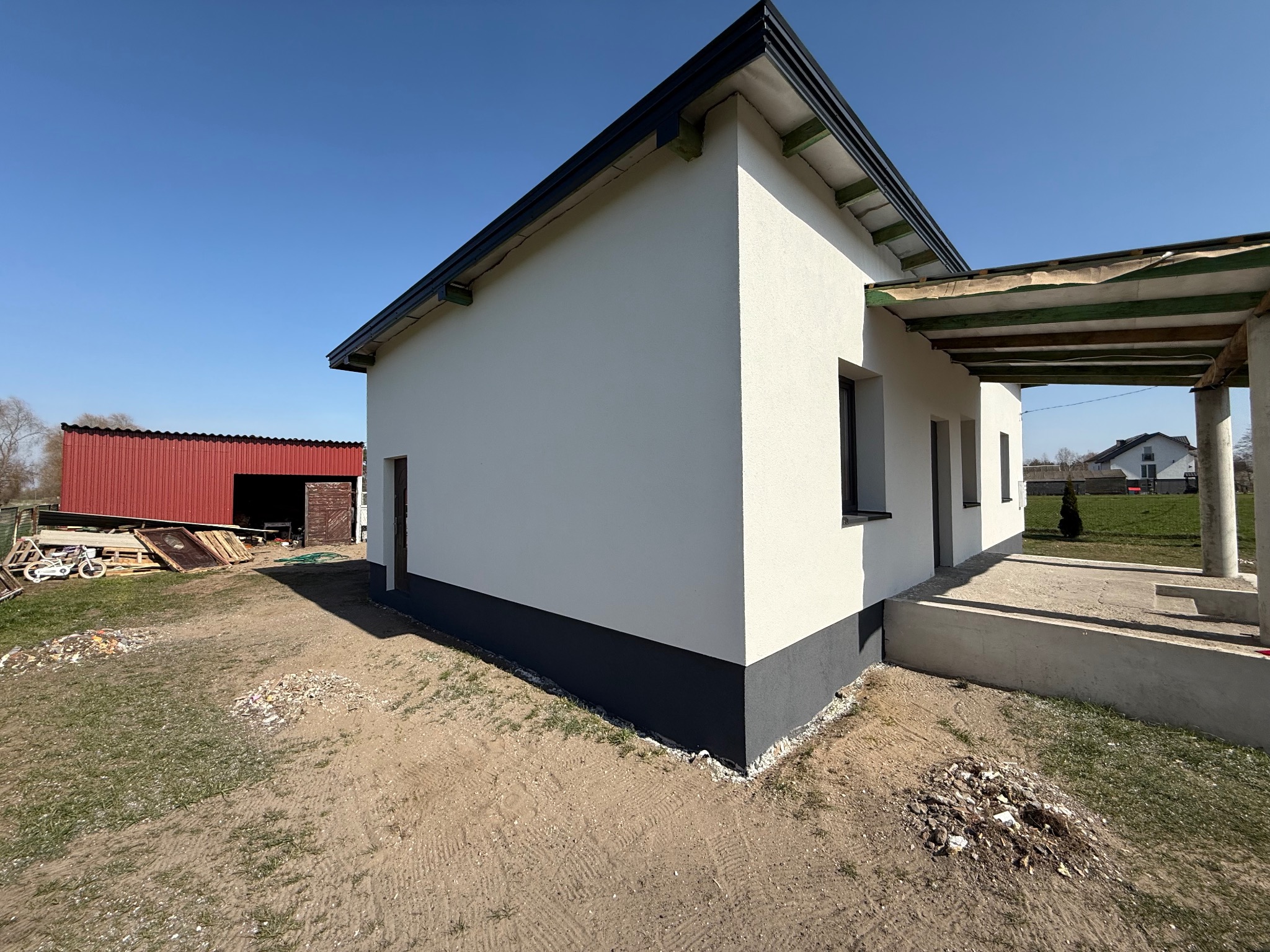 Boczny widok otynkowanego domu z ciemną podmurówką i fragmentem zadaszonego tarasu w trakcie wykończenia, na tle błękitnego nieba i zabudowań gospodarczych.