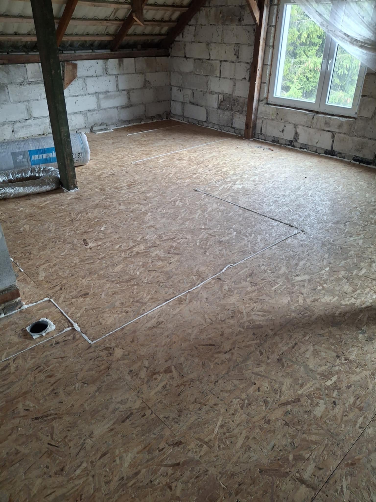 Płyty OSB na poddaszu, przygotowane do dalszych prac wykończeniowych. Widoczne białe uszczelnienia i izolacja. Surowy stan, okno z widokiem na zieleń.