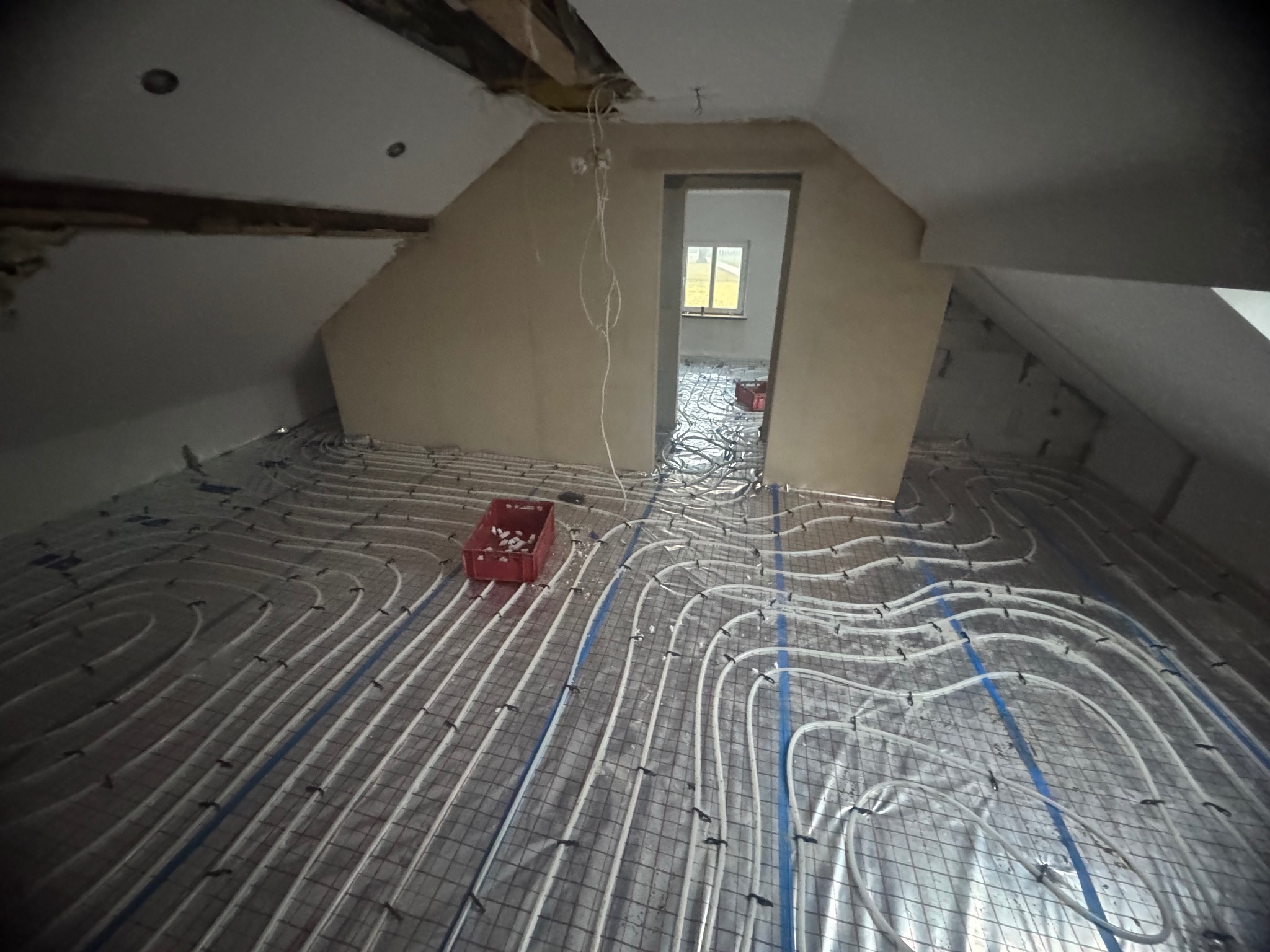 Instalacja ogrzewania podłogowego w surowym wnętrzu poddasza. Białe rury ułożone na siatce, widoczne elementy konstrukcyjne dachu i okno w oddali. Czerwony pojemnik na podłodze.