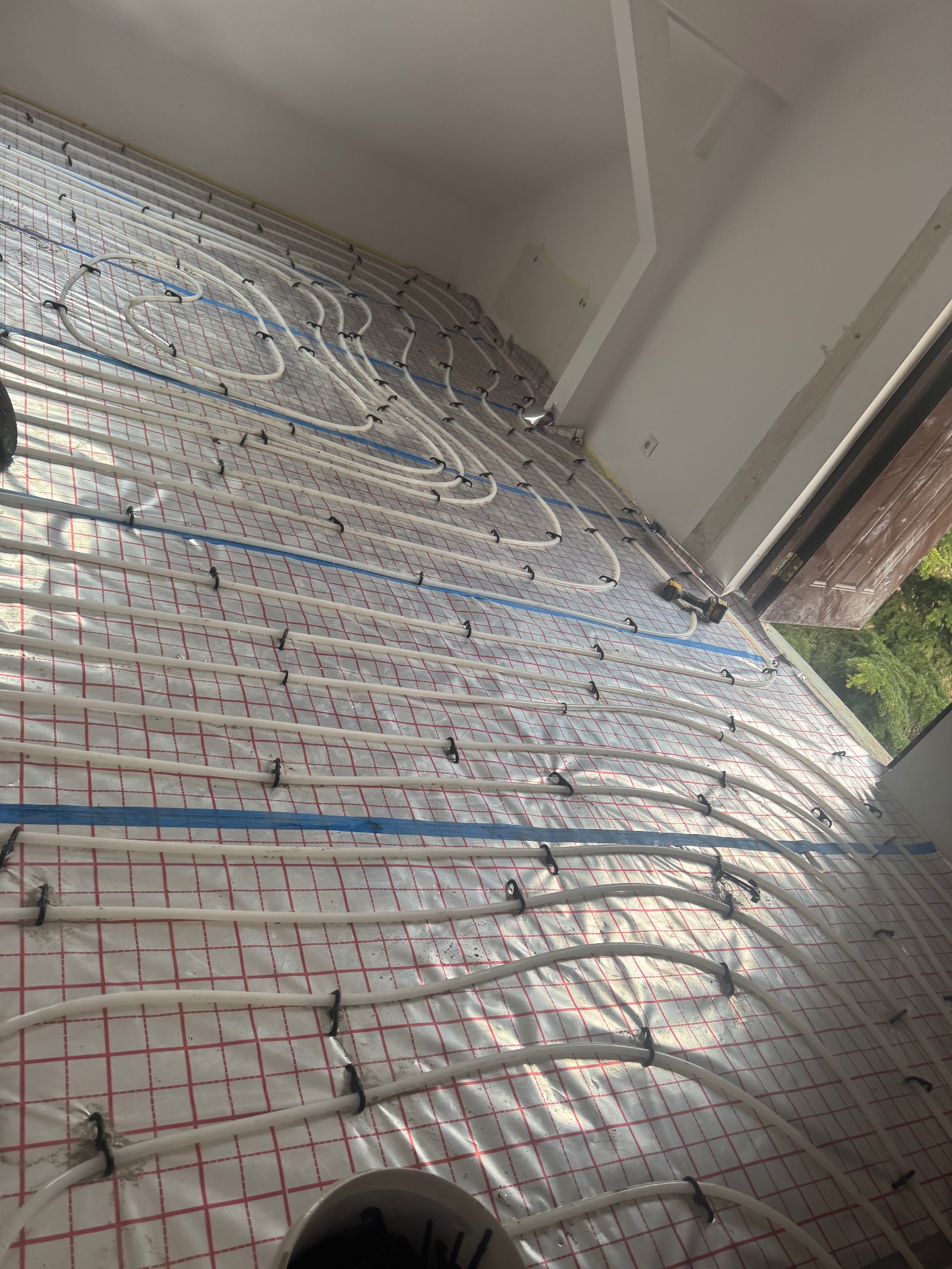 Białe rury ogrzewania podłogowego, przymocowane czarnymi uchwytami do siatki na izolacji, ułożone w jasnym pomieszczeniu. Widoczne narzędzia.