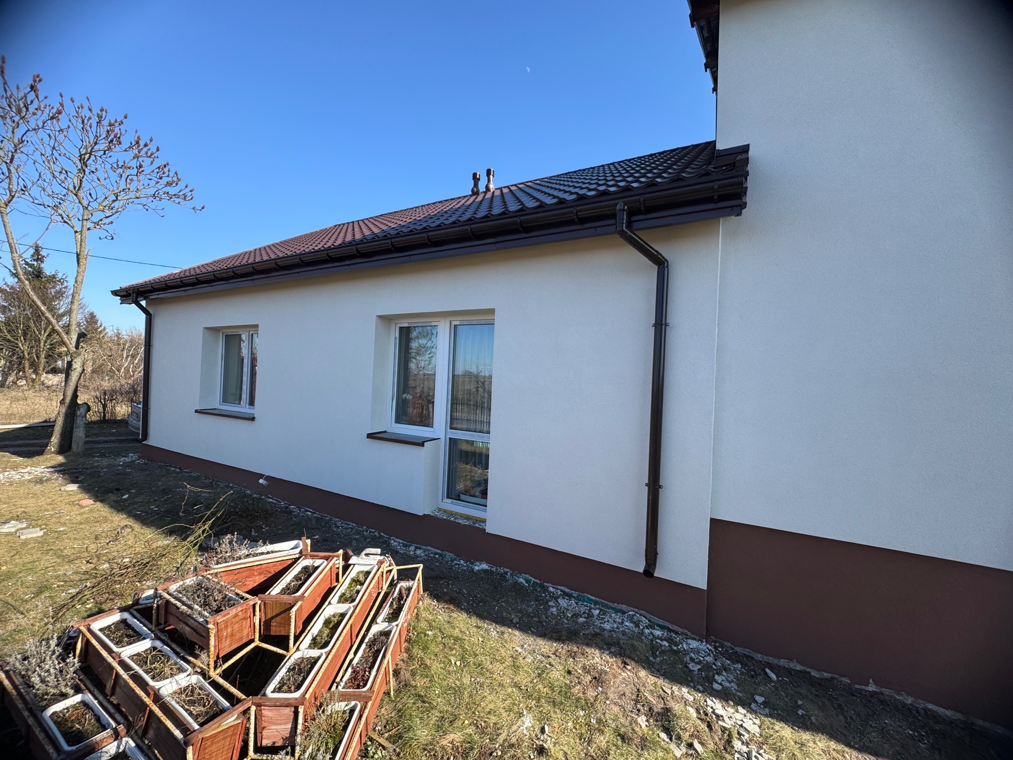 Otynkowana, jasna elewacja domu jednorodzinnego z brązowym dachem i rynnami, widoczna z boku w słoneczny dzień. W tle drzewo bez liści i błękitne niebo.