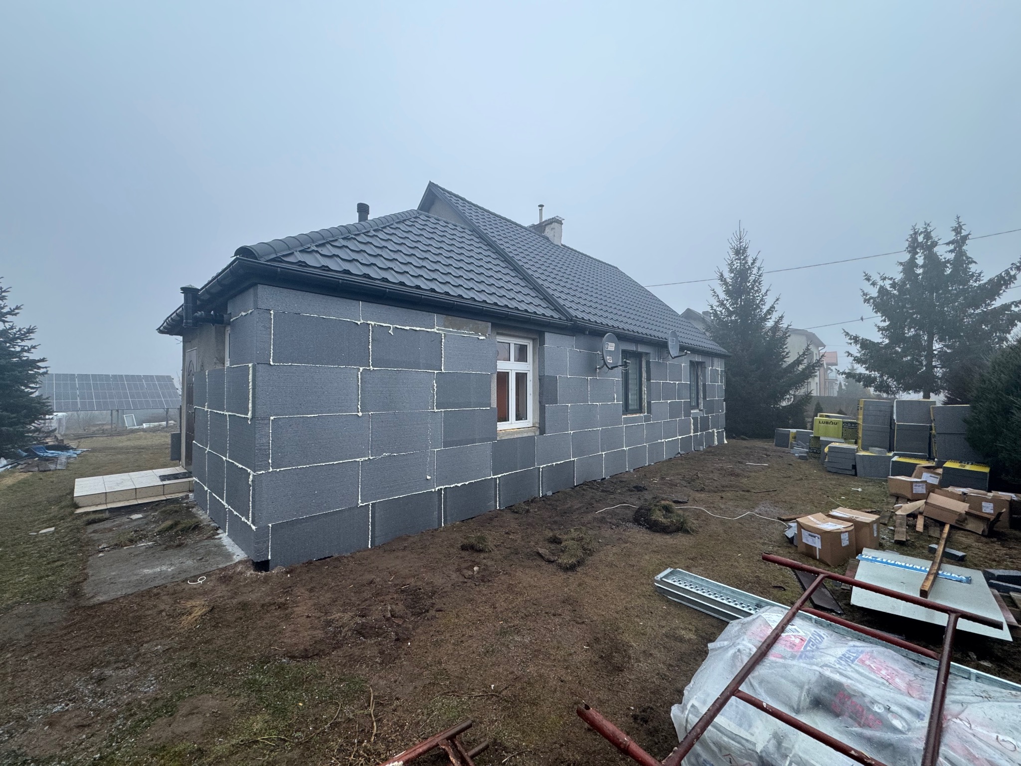 Dom jednorodzinny w trakcie ocieplania, ściany pokryte szarymi płytami izolacyjnymi. Widoczne okna, dach z ciemnej dachówki i elementy rusztowania na posesji.