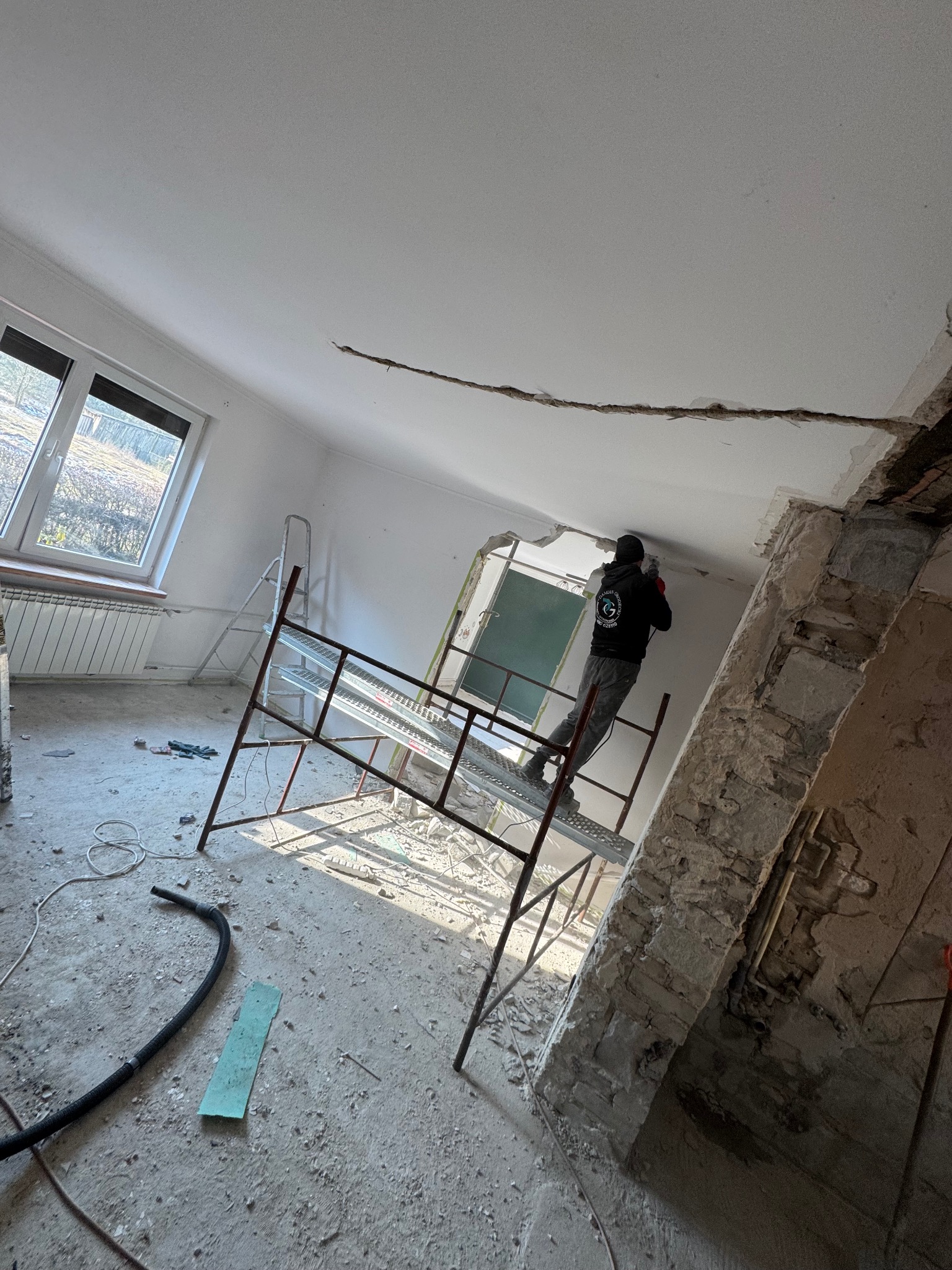 Pracownik na rusztowaniu skuwa fragment sufitu w jasnym pomieszczeniu z oknem. Widoczne gruz i narzędzia, co sugeruje etap remontu domu lub kamienicy.