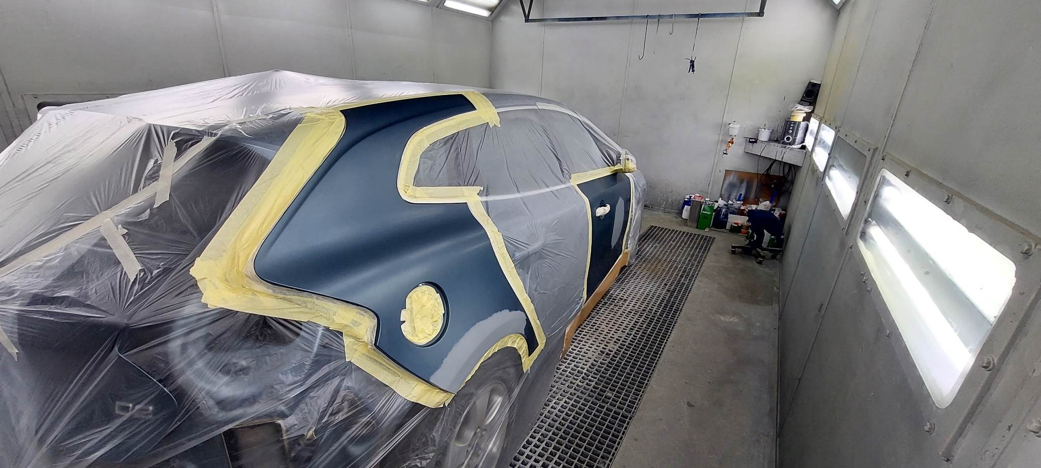 Przygotowanie auta do lakierowania w kabinie: oklejone taśmą i folią elementy karoserii w kolorze granatowym. Widoczny fragment felgi i kratka ściekowa.