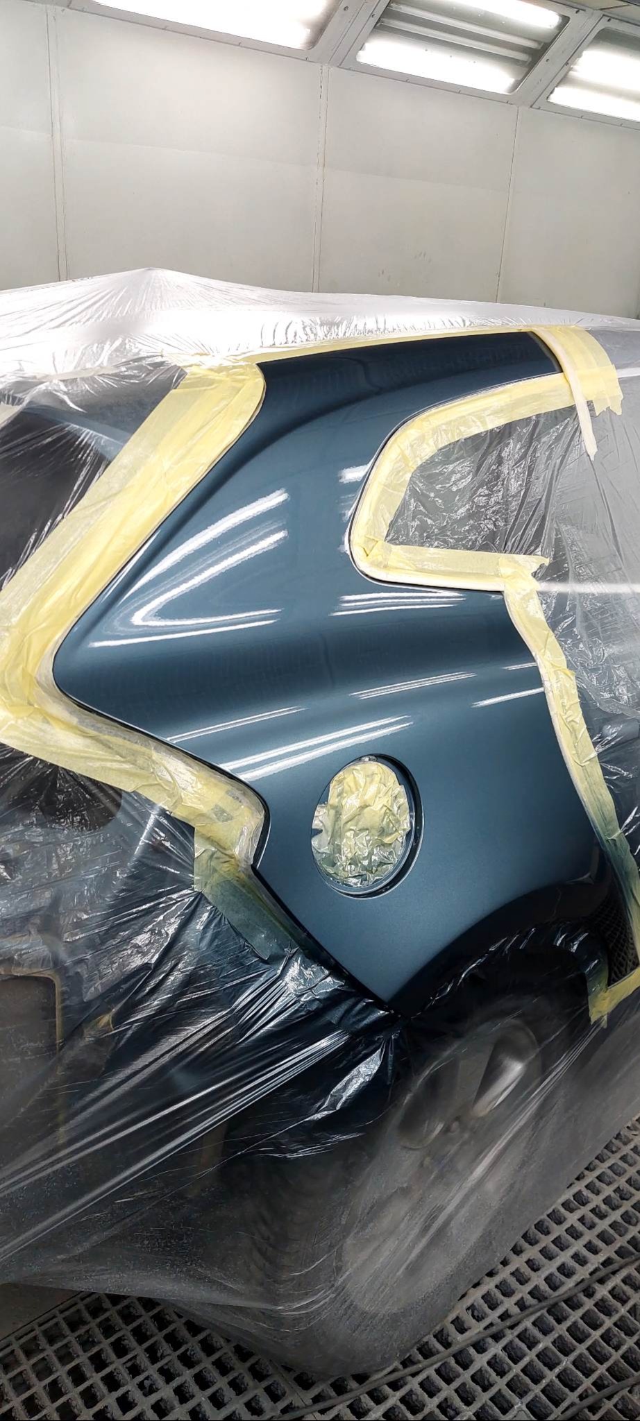 Boczny fragment karoserii auta w odcieniach szarości, zabezpieczony folią i taśmą malarską w kabinie lakierniczej, przygotowany do lakierowania. Widoczne refleksy światła.