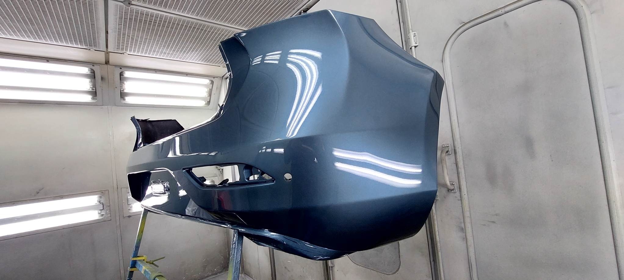 Pomalowany na szaro tylny zderzak samochodu w kabinie lakierniczej, z widocznymi refleksami światła na gładkiej powierzchni. Zderzak umieszczony na stojaku.