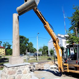 NASTES KAMIL CHILIMONIUK - Żółta koparka umieszcza element kolumny na kamiennej podstawie. W tle budynki i drzewa w słoneczny dzień. Etap prac budowlanych.