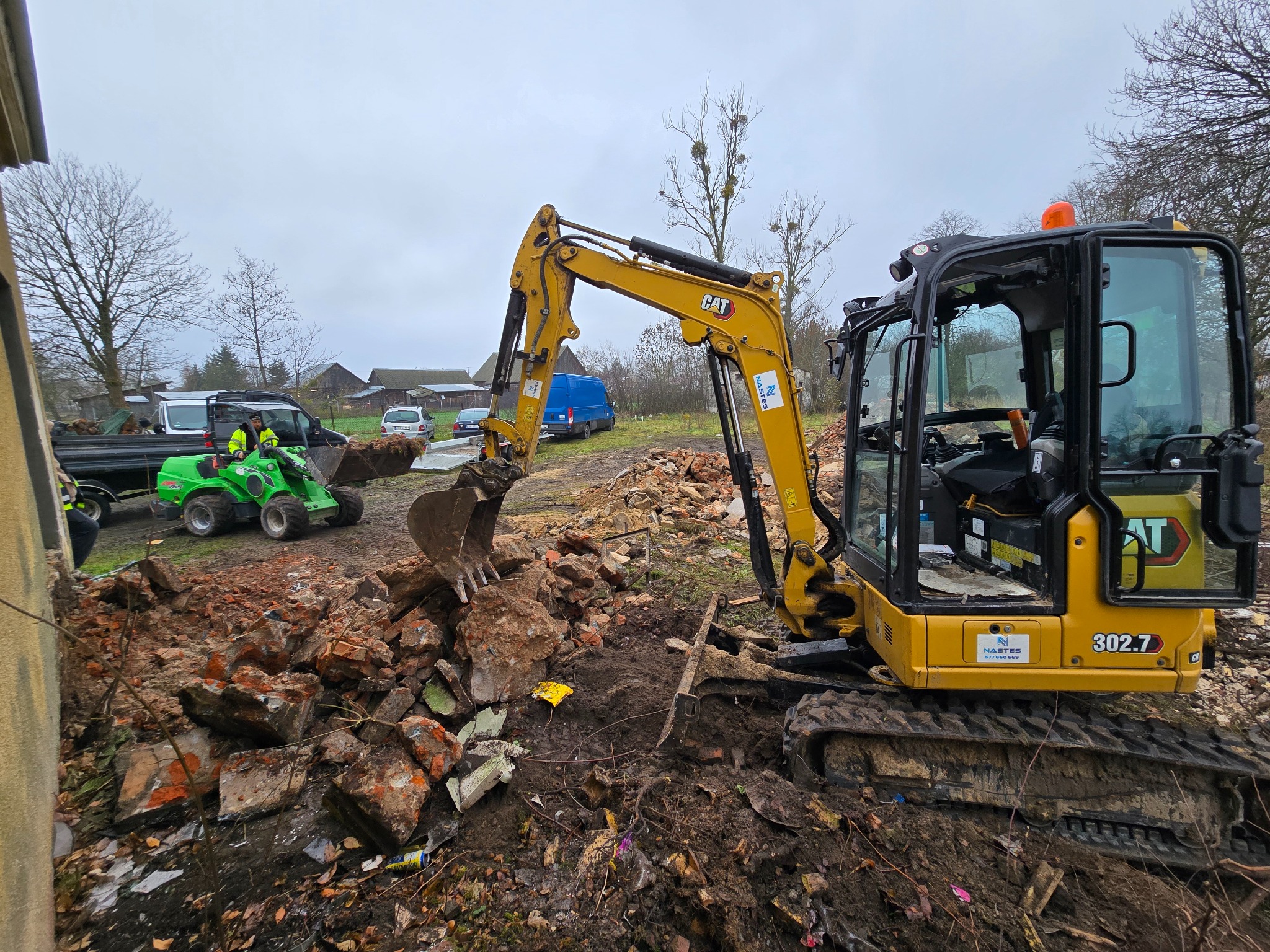 Żółta mini koparka CAT 302.7 w trakcie prac rozbiórkowych, gruz ceglany wokół, w tle zielony traktor i budynki gospodarcze, pochmurne niebo.