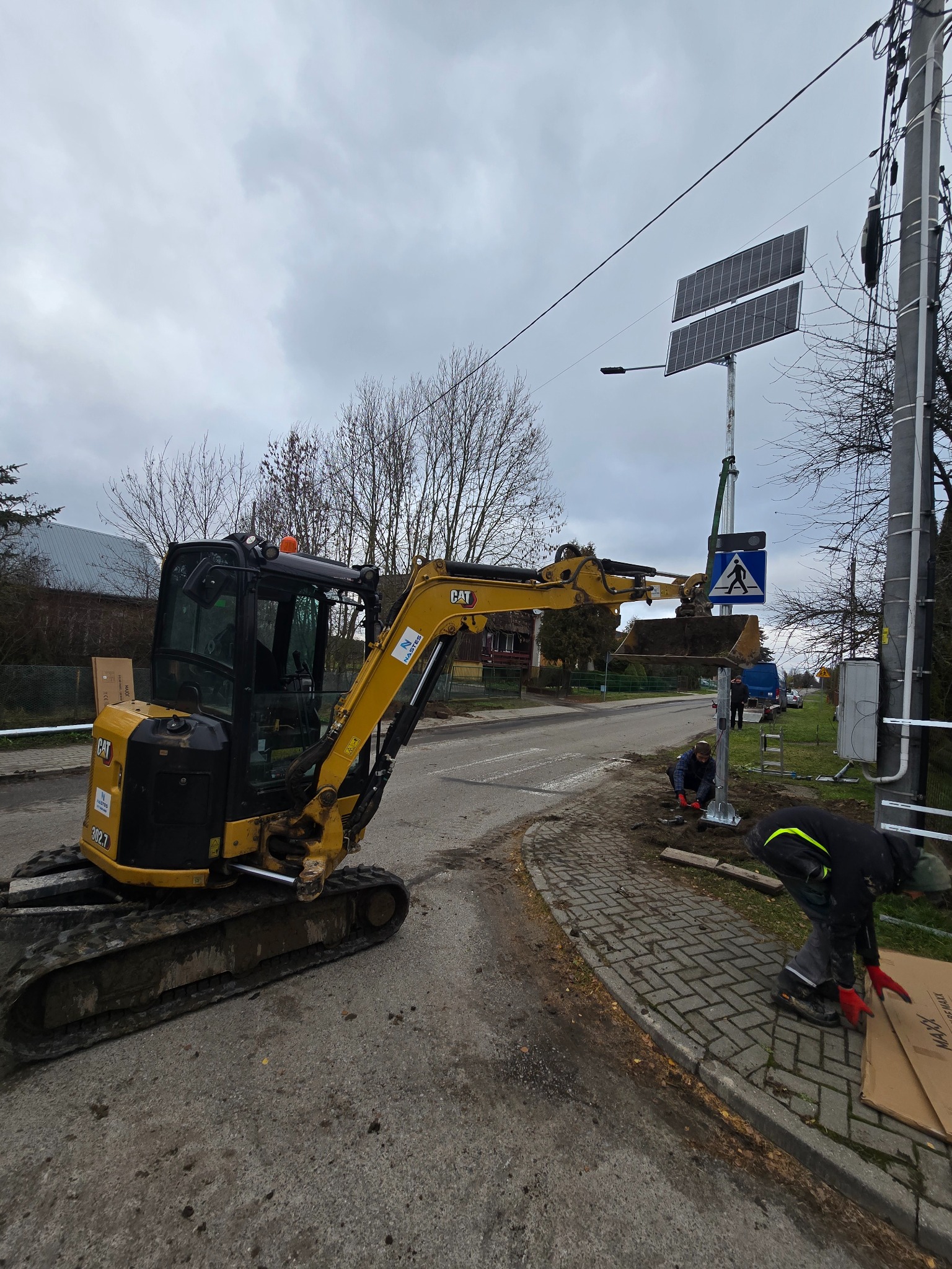 Żółta mini koparka CAT 302.7 przy montażu znaku przejścia dla pieszych zasilanego panelami słonecznymi. Dwóch pracowników przy pracy, kostka brukowa, pochmurne niebo.