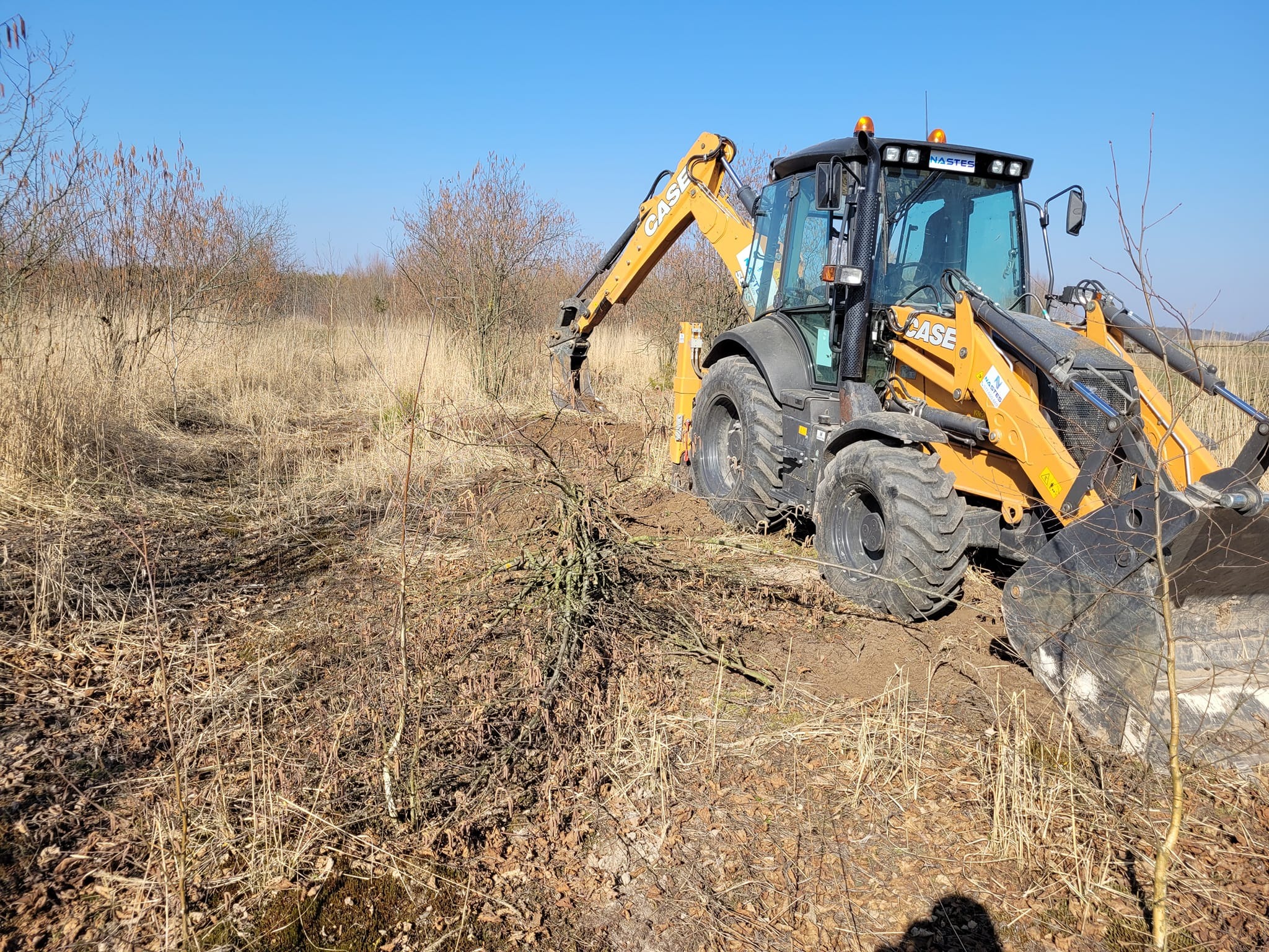 Żółta koparko-ładowarka Case w trakcie prac ziemnych na zarośniętym terenie w słoneczny dzień. Widoczny wykop i resztki roślinności.