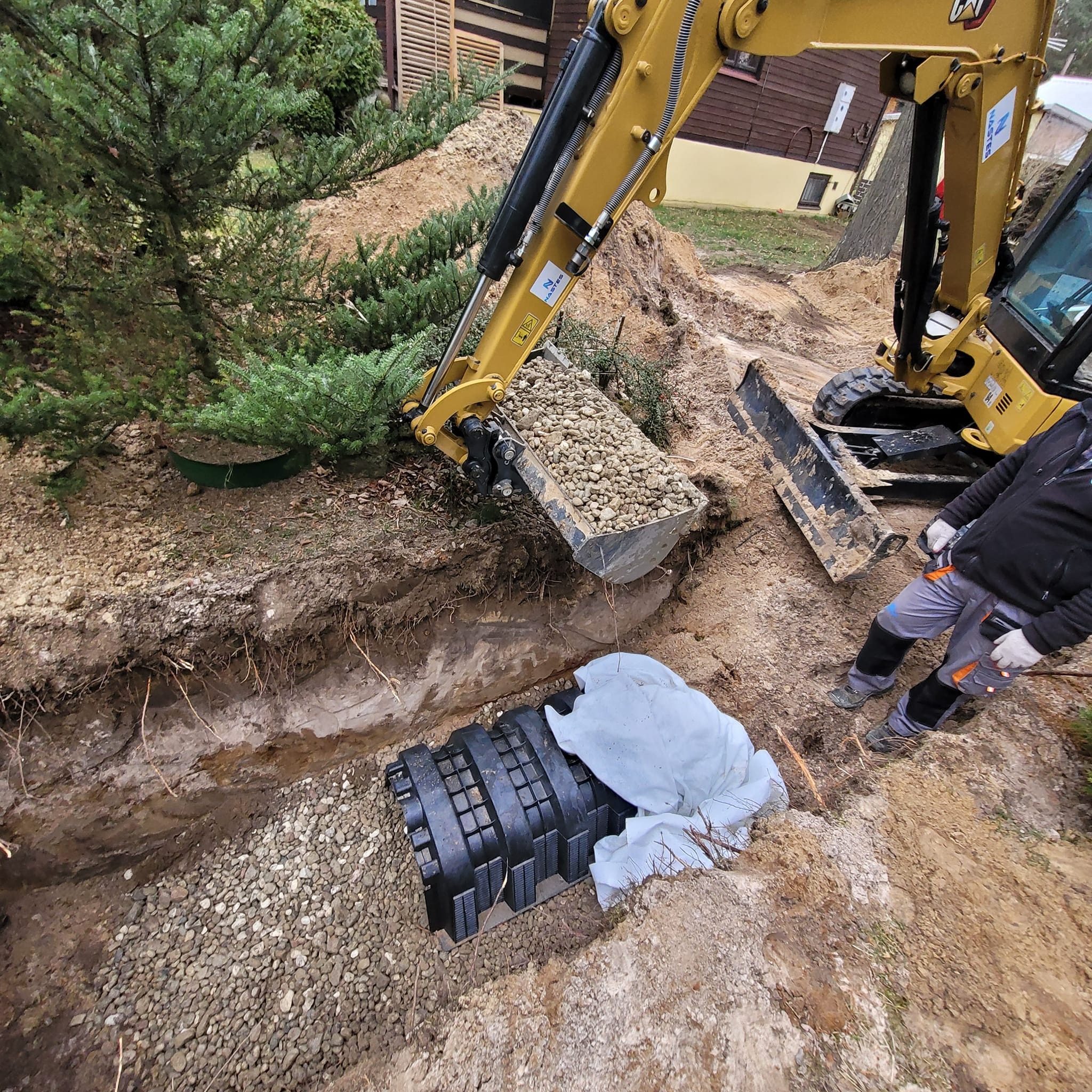 Żółta koparka zasypuje żwirem czarny zbiornik w wykopie. Widoczny pracownik w szarych spodniach roboczych. W tle iglaste drzewo i budynek.