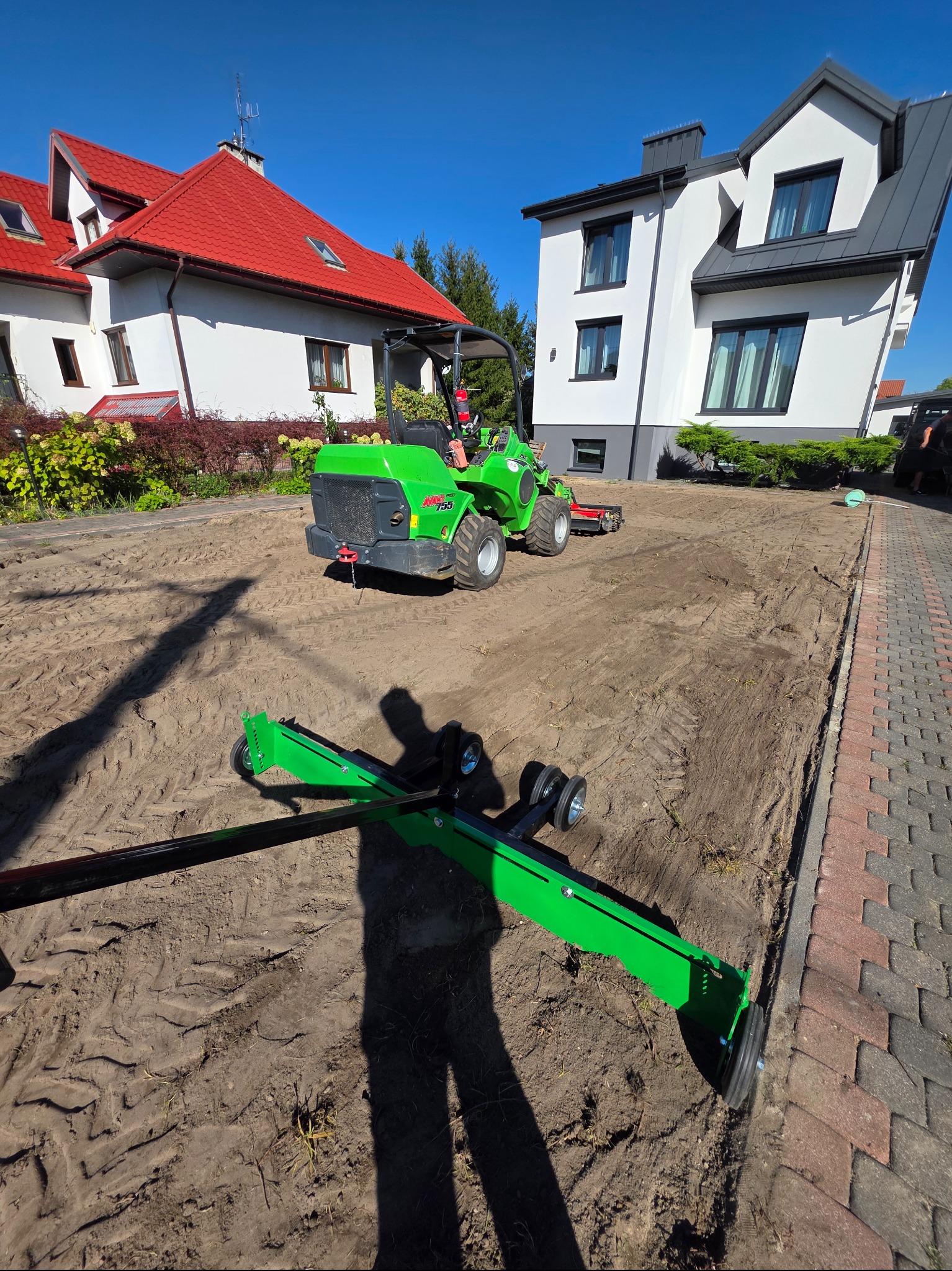 Przygotowanie terenu pod trawnik: miniładowarka Avant 755 z glebogryzarką wyrównuje ziemię na działce. W tle domy z czerwoną i szarą dachówką, błękitne niebo.