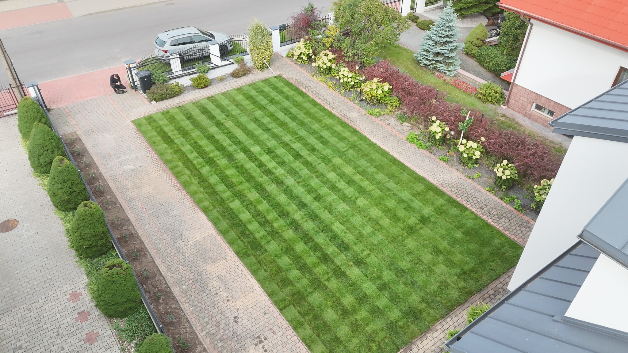 Z góry: Idealnie przystrzyżony trawnik w pasy, obramowany kostką brukową i zielenią. Osoba przy bramie, auto na podjeździe, zadbany ogród z kwiatami i krzewami.