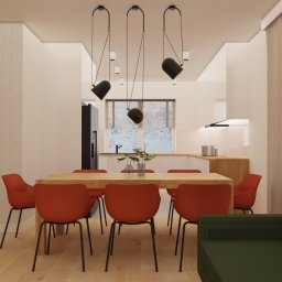 Jasne wnętrze z drewnianym stołem i czerwonymi krzesłami. W tle widoczna kuchnia z oknem na zimowy krajobraz. Minimalistyczne lampy wiszące nad stołem.