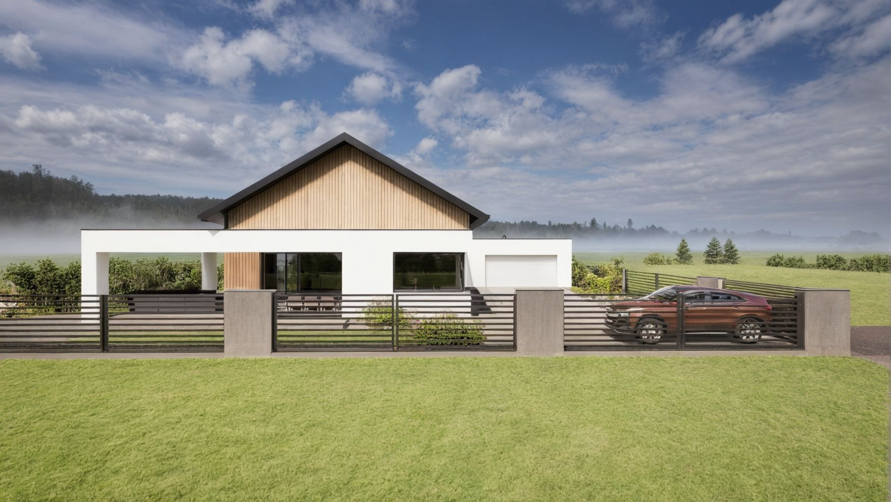 Nowoczesny dom jednorodzinny z drewnianą elewacją i dużymi oknami, otoczony zielonym trawnikiem i ogrodzeniem. Na podjeździe stoi bordowy samochód. W tle mglista łąka.
