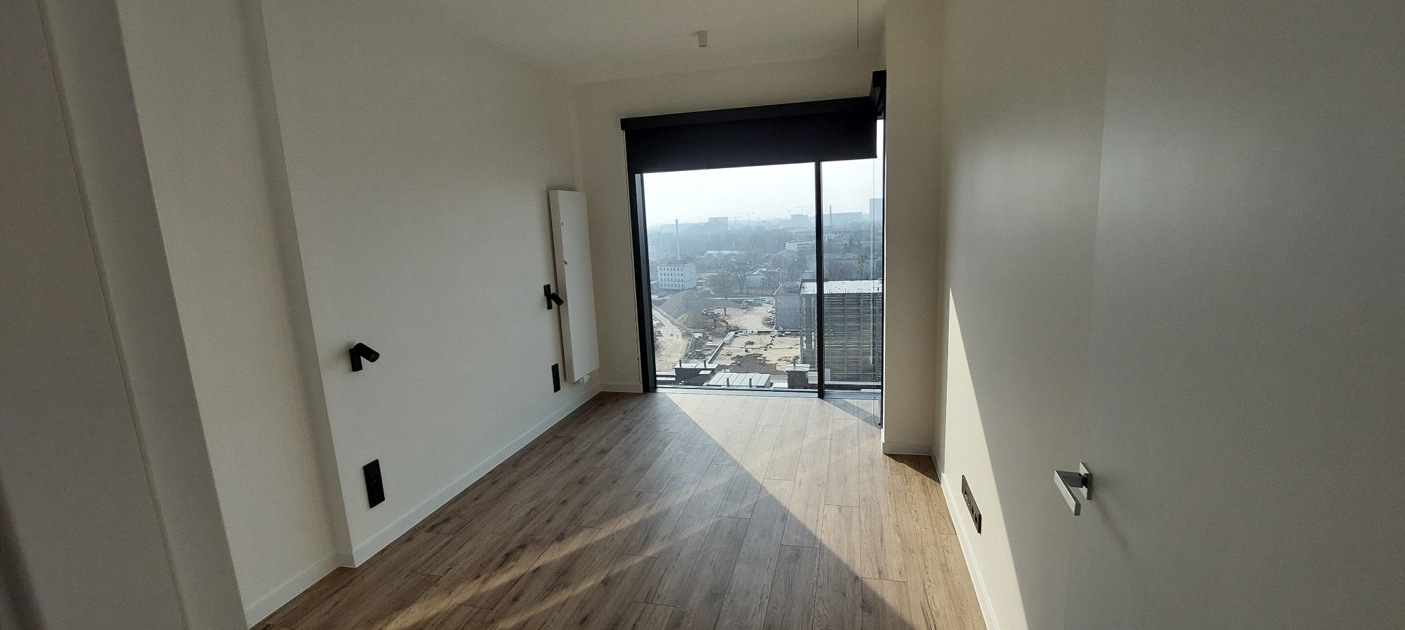 Jasne wnętrze z drewnopodobną podłogą i dużym oknem z widokiem na miasto. Na ścianie minimalistyczny grzejnik i czarne punkty oświetleniowe.