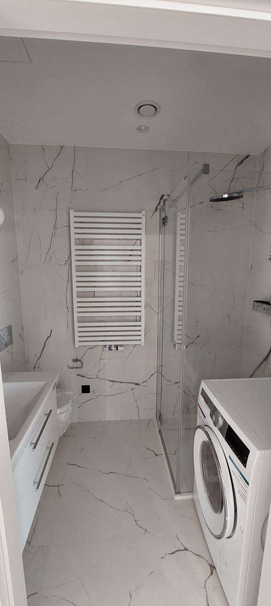 Jasna łazienka z marmurowymi płytkami, kabiną prysznicową, grzejnikiem drabinkowym i pralką. Minimalistyczny design, nowoczesne wykończenie.