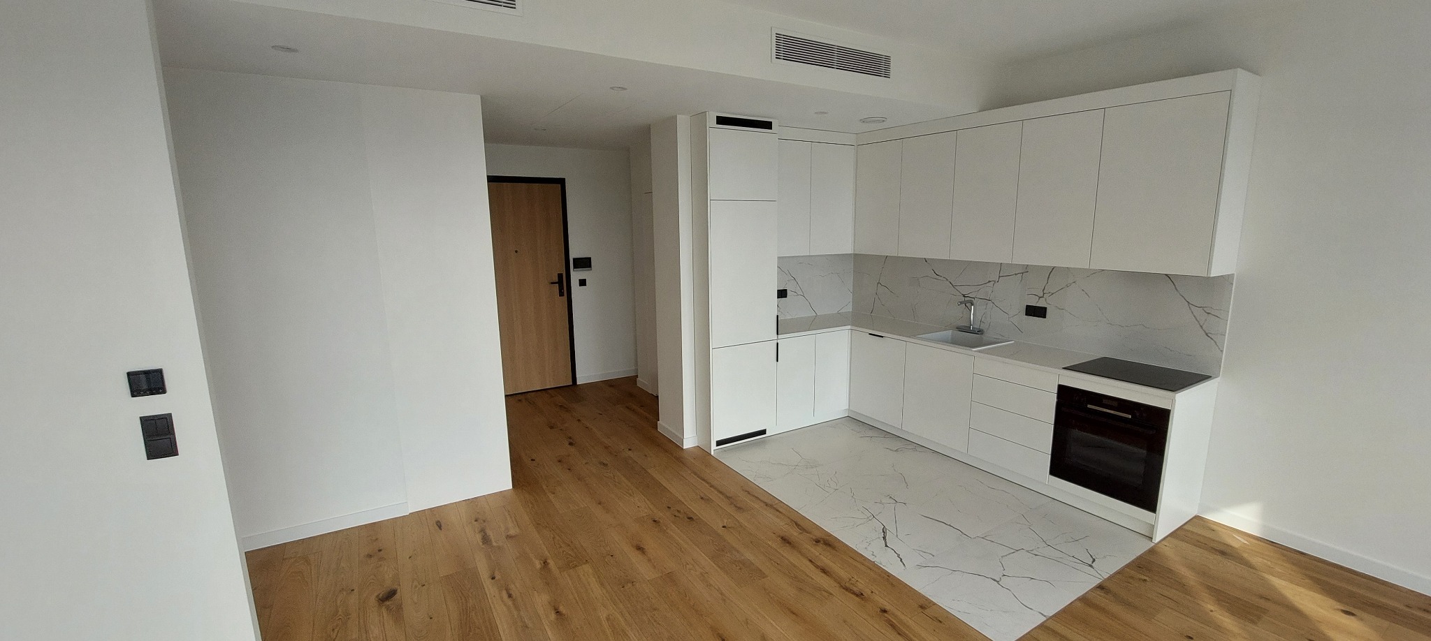 Nowoczesna, jasna kuchnia z białymi szafkami i blatem imitującym marmur. Drewniana podłoga przechodzi w marmurową. Widoczne minimalistyczne detale i czarne akcenty.