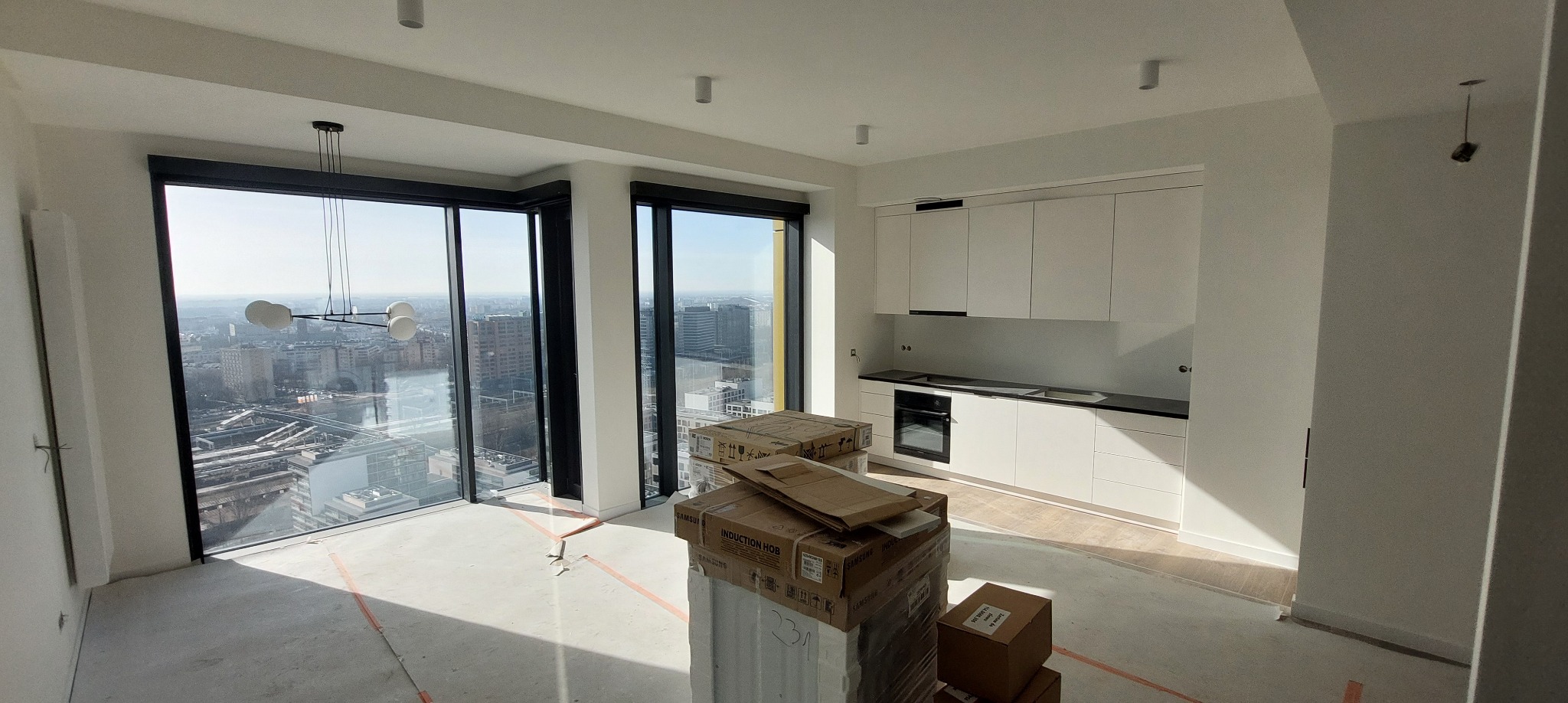 Wnętrze apartamentu w trakcie wykończenia: biała zabudowa kuchenna, duże okna z widokiem na miasto, kartony z nowym sprzętem AGD. Surowa podłoga przed położeniem paneli.