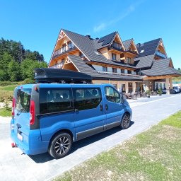 BIKE-BUS KAMIL MATUSIAK - Niebieski bus z bagażnikiem dachowym na tle drewnianego hotelu w górach, błękitne niebo. Transport osób i bagażu w malowniczej scenerii.