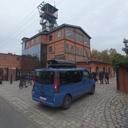 BIKE-BUS KAMIL MATUSIAK - Niebieski bus Renault Trafic z bagażnikiem dachowym na tle zabytkowej kopalni 'Szyb Maciej'. Pojazd zaparkowany na brukowanej drodze, w tle spacerujący ludzie.